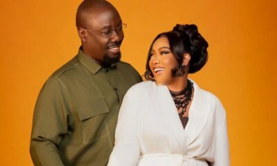 Couple smiling at each other against an orange background; man in an olive shirt, woman in a white blazer with black jewelry.