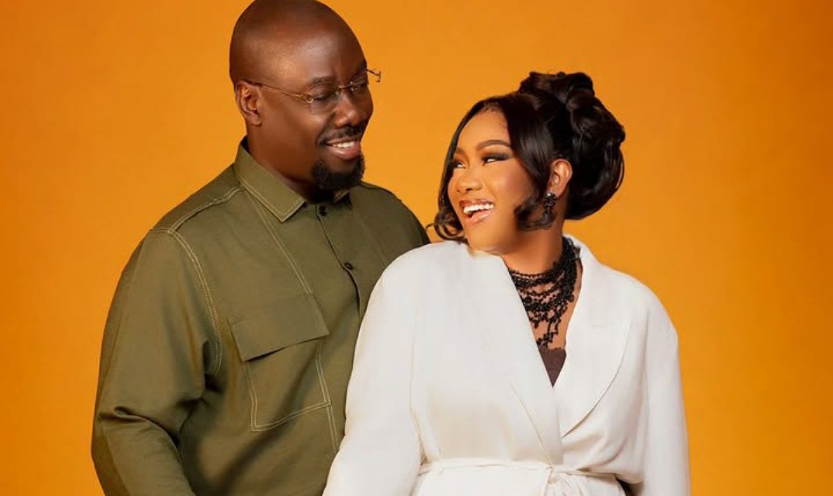 Couple smiling at each other against an orange background; man in an olive shirt, woman in a white blazer with black jewelry.