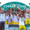 Group of young athletes in white shirts and yellow pants celebrate on a podium with trophies and medals, champion banner overhead reads 'CHAMPIONS'.