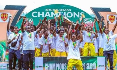 Group of young athletes in white shirts and yellow pants celebrate on a podium with trophies and medals, champion banner overhead reads 'CHAMPIONS'.