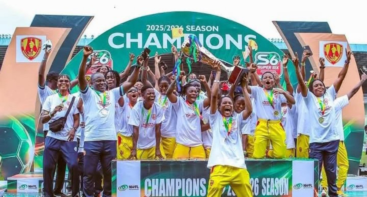 Group of young athletes in white shirts and yellow pants celebrate on a podium with trophies and medals, champion banner overhead reads 'CHAMPIONS'.