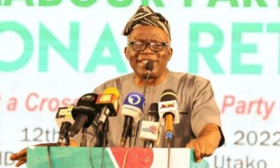 Speaker at a press conference wearing a traditional hat and glasses, standing behind a colorful podium with multiple microphones from different media outlets.