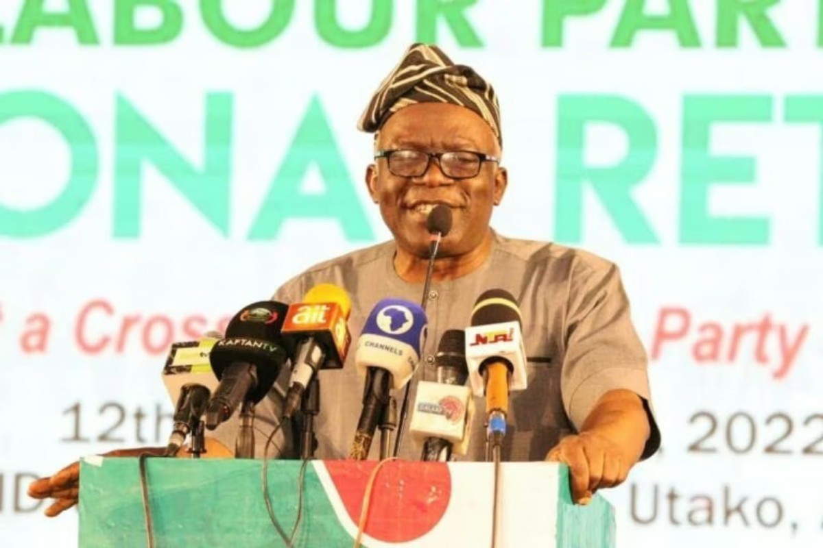 Speaker at a press conference wearing a traditional hat and glasses, standing behind a colorful podium with multiple microphones from different media outlets.