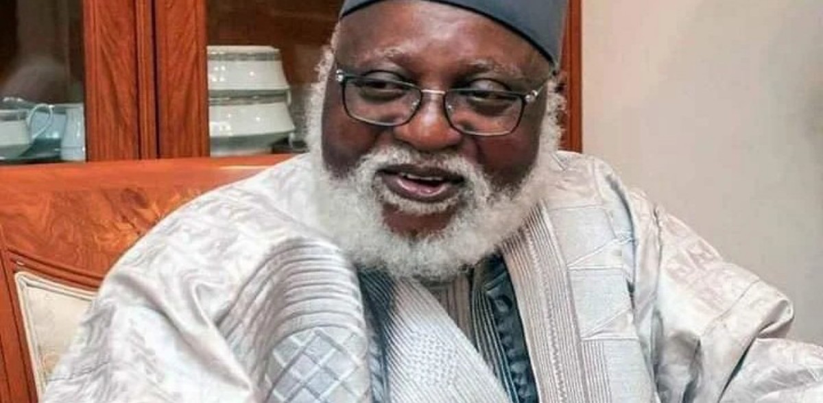 Smiling older man with a white beard and glasses, wearing traditional gray attire and a cap, indoors.