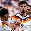 German national team players celebrate a goal, wearing white jerseys with black-red-gold stripes; Musiala visible on the back of one jersey.