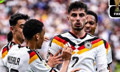 German national team players celebrate a goal, wearing white jerseys with black-red-gold stripes; Musiala visible on the back of one jersey.
