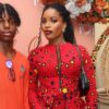 Two models posing at a fashion event: the woman in a red star-pattern dress with a black bag, and the man in an orange shirt against a pink backdrop.