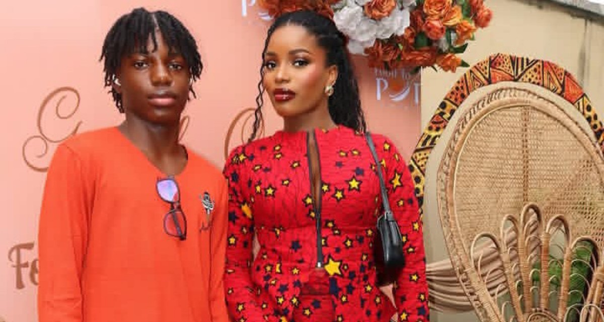 Two models posing at a fashion event: the woman in a red star-pattern dress with a black bag, and the man in an orange shirt against a pink backdrop.