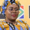 Portrait of a woman wearing a blue and yellow patterned head wrap and matching outfit, with glasses, speaking at an event.