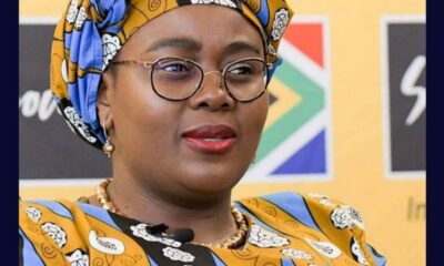 Portrait of a woman wearing a blue and yellow patterned head wrap and matching outfit, with glasses, speaking at an event.