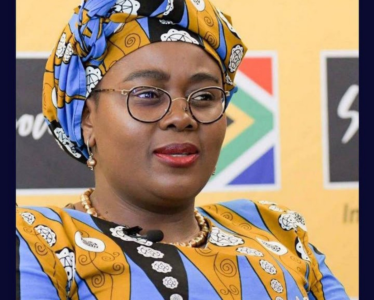 Portrait of a woman wearing a blue and yellow patterned head wrap and matching outfit, with glasses, speaking at an event.