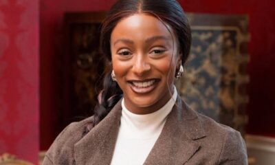 Smiling woman in a brown blazer and white turtleneck, posing indoors with a warm, elegant backdrop.