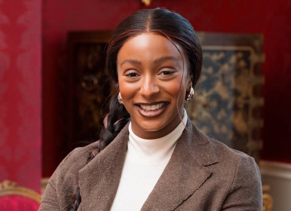 Smiling woman in a brown blazer and white turtleneck, posing indoors with a warm, elegant backdrop.
