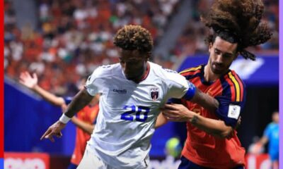 Two soccer players duel for the ball during a match; one in a white kit with number 28, the other in a red-and-blue kit with long hair tied back. Crowded stands in the background.