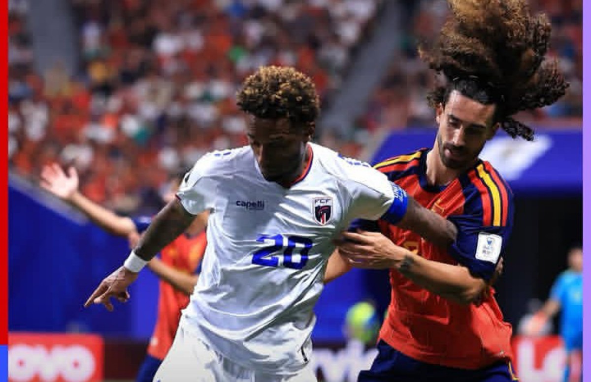 Two soccer players duel for the ball during a match; one in a white kit with number 28, the other in a red-and-blue kit with long hair tied back. Crowded stands in the background.