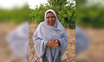 Woman in a light gray hijab stands in a dry, grassy field with green bushes in the background, hands folded.