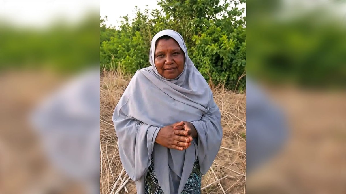 Woman in a light gray hijab stands in a dry, grassy field with green bushes in the background, hands folded.