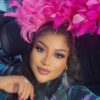 Close-up portrait of a woman wearing a bright pink feather headpiece inside a car, with bold makeup and a gentle smile.