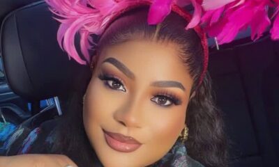 Close-up portrait of a woman wearing a bright pink feather headpiece inside a car, with bold makeup and a gentle smile.