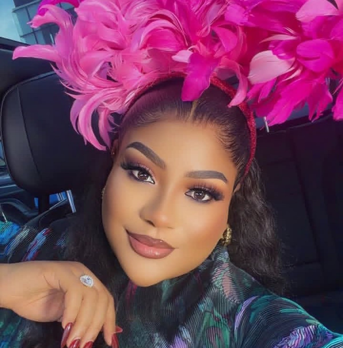 Close-up portrait of a woman wearing a bright pink feather headpiece inside a car, with bold makeup and a gentle smile.