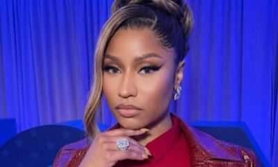 Woman posing with hand under chin, wearing a red shirt and textured burgundy jacket, with glamorous makeup and a blue backdrop