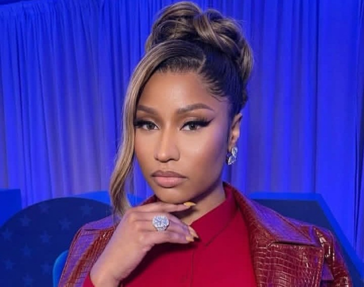 Woman posing with hand under chin, wearing a red shirt and textured burgundy jacket, with glamorous makeup and a blue backdrop