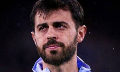 Close-up of a man with dark hair and beard, wearing a light blue zip-up jacket, looking to the side against a dark background.