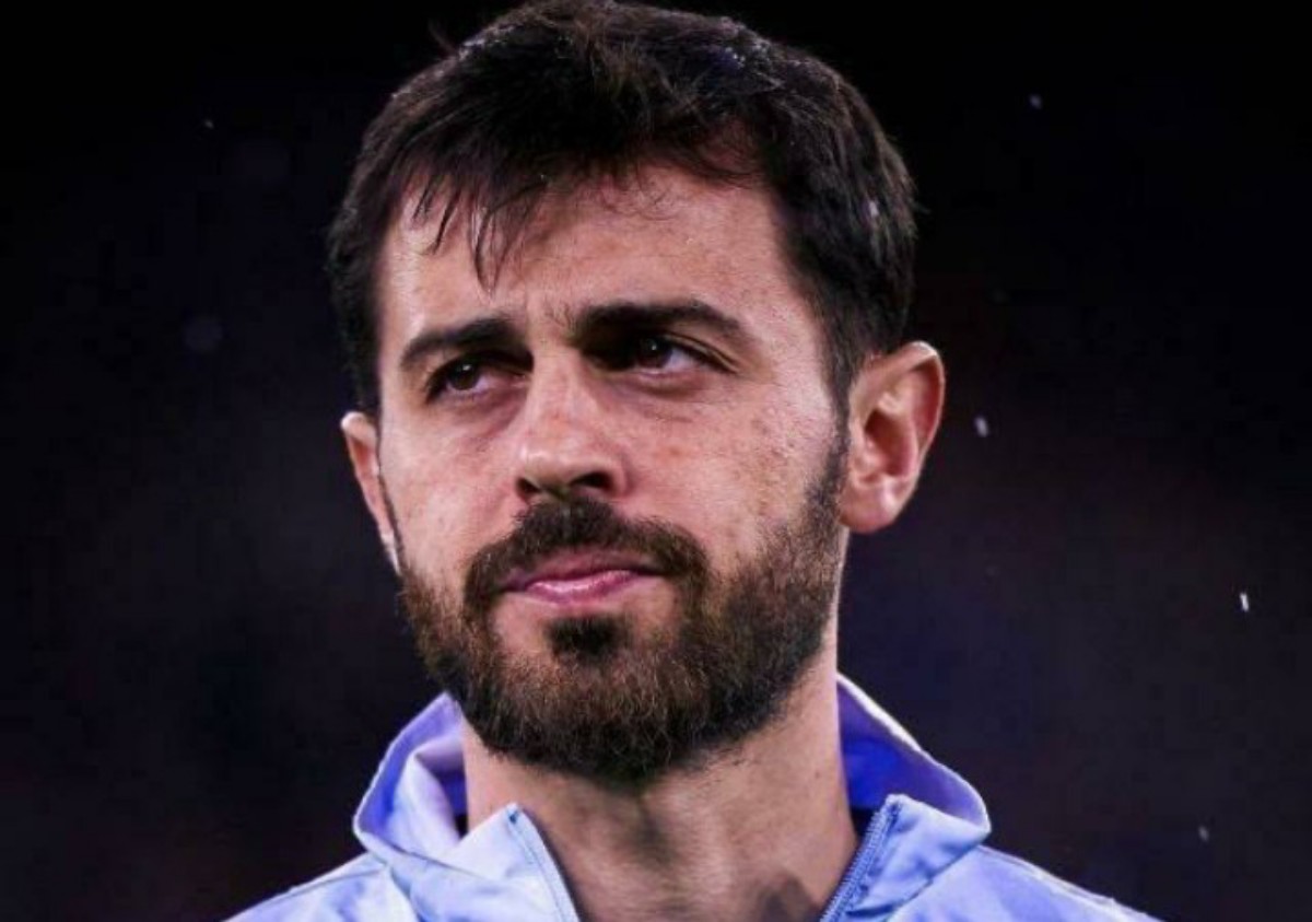 Close-up of a man with dark hair and beard, wearing a light blue zip-up jacket, looking to the side against a dark background.
