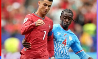 Two soccer players, one in a red kit and one in a light blue kit, contest for the ball during a match in a crowded stadium.