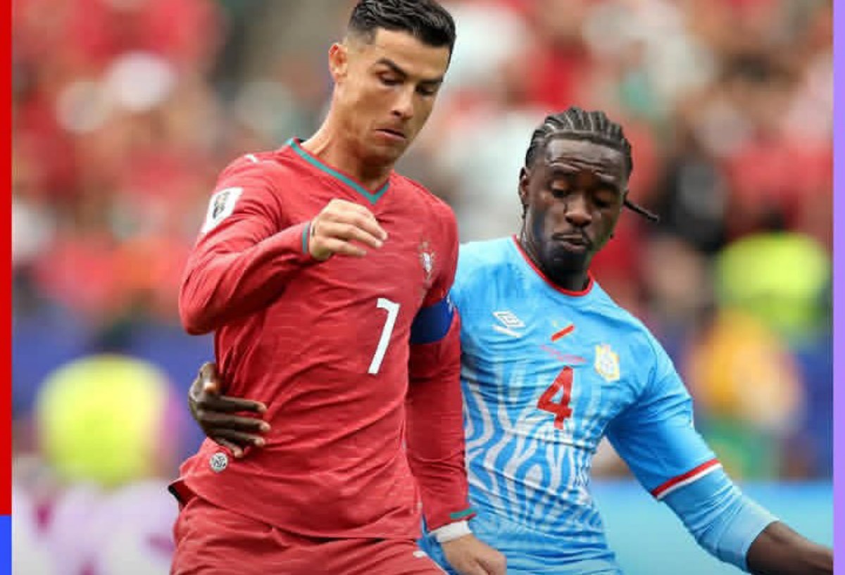 Two soccer players, one in a red kit and one in a light blue kit, contest for the ball during a match in a crowded stadium.