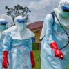 Line of health workers in full protective gear outdoors, wearing blue suits, red gloves, and face shields, with a spray device in use.