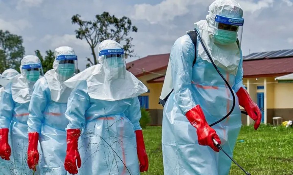 Line of health workers in full protective gear outdoors, wearing blue suits, red gloves, and face shields, with a spray device in use.