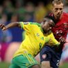Two soccer players clash for the ball during a match: one in a yellow Brazil kit, the other in red (Wales) kit, in a blurred stadium background.