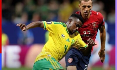 Two soccer players clash for the ball during a match: one in a yellow Brazil kit, the other in red (Wales) kit, in a blurred stadium background.