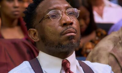 Close-up of a man in round glasses, white shirt, red patterned tie and suspenders, seated in an audience and looking upward thoughtfully.