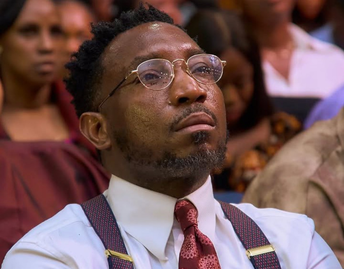 Close-up of a man in round glasses, white shirt, red patterned tie and suspenders, seated in an audience and looking upward thoughtfully.