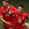 Two soccer players in red uniforms celebrate together on a green pitch, one with a raised hand.