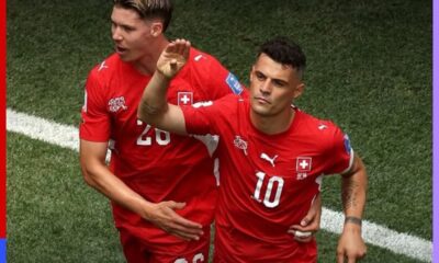 Two soccer players in red uniforms celebrate together on a green pitch, one with a raised hand.
