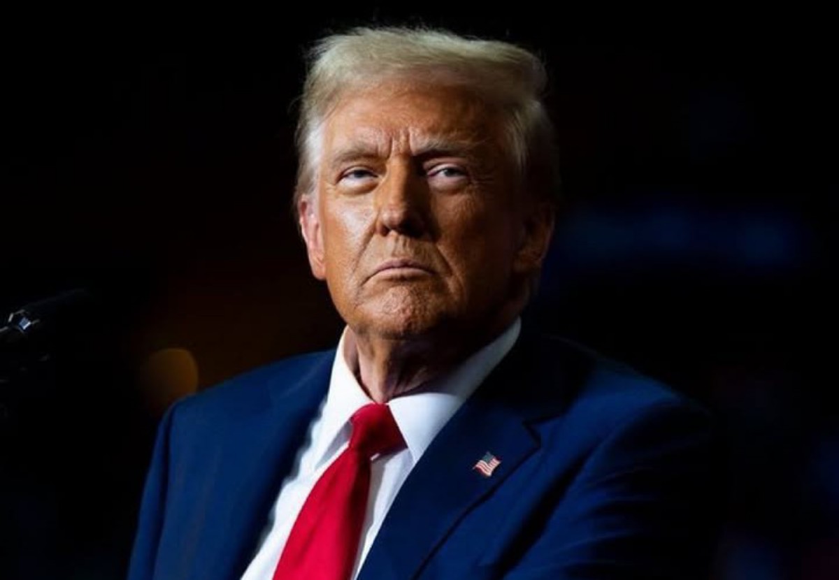 Solemn man in a dark suit with a red tie and American flag pin, against a dark background.
