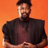 Portrait of a man with short dreadlocks and a goatee wearing a black traditional outfit against an orange background, hands clasped in front.