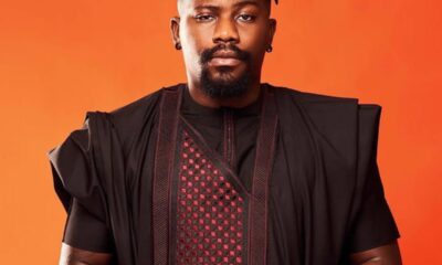 Portrait of a man with short dreadlocks and a goatee wearing a black traditional outfit against an orange background, hands clasped in front.