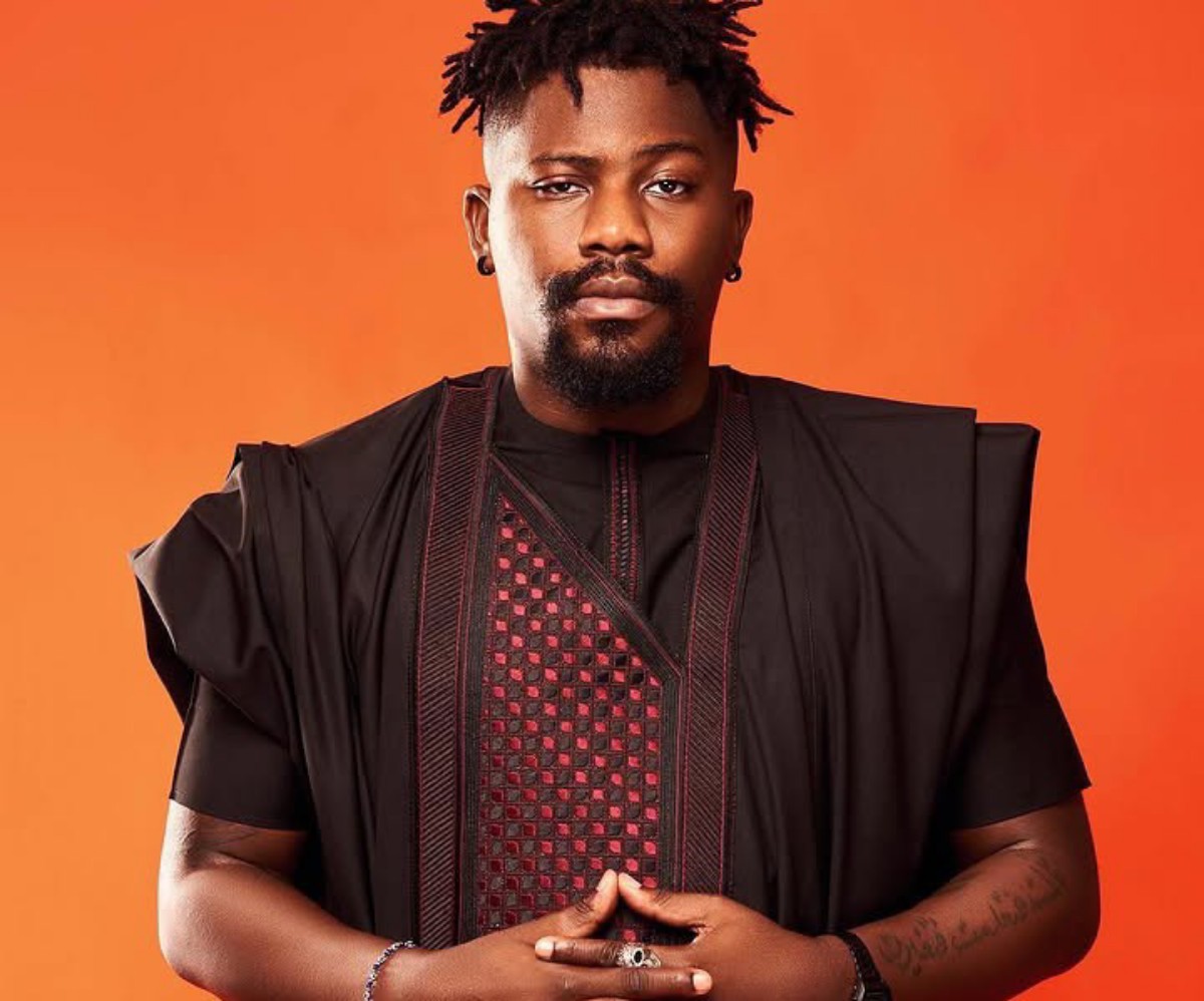 Portrait of a man with short dreadlocks and a goatee wearing a black traditional outfit against an orange background, hands clasped in front.