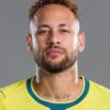Close-up portrait of a man with curly hair and beard wearing a yellow sports jersey against a gray background, visible neck tattoos.