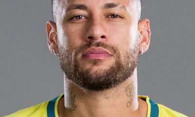 Close-up portrait of a man with curly hair and beard wearing a yellow sports jersey against a gray background, visible neck tattoos.