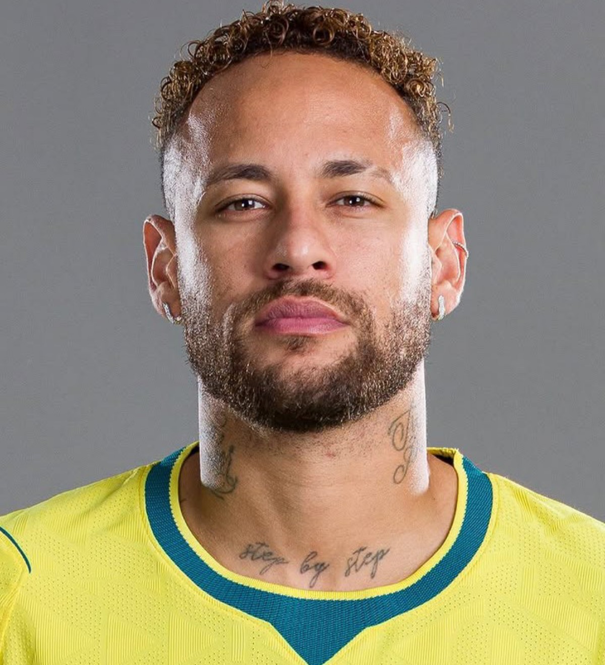 Close-up portrait of a man with curly hair and beard wearing a yellow sports jersey against a gray background, visible neck tattoos.