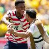 Two USA soccer players celebrate on the field, one in a red-and-white striped jersey with number 16, smiling broadly with arms out.