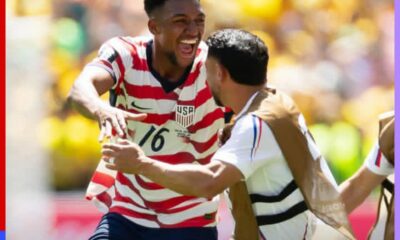 Two USA soccer players celebrate on the field, one in a red-and-white striped jersey with number 16, smiling broadly with arms out.