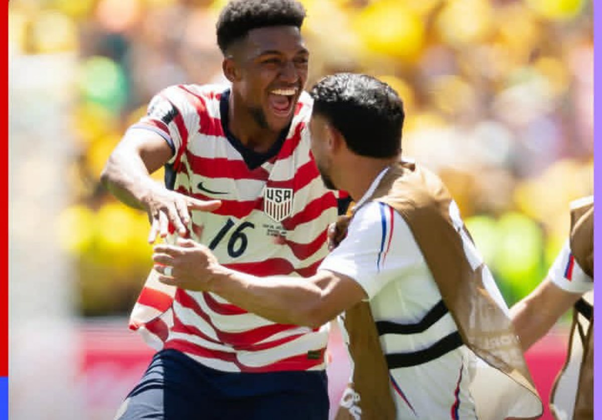 Two USA soccer players celebrate on the field, one in a red-and-white striped jersey with number 16, smiling broadly with arms out.
