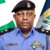 Portrait of a male police officer in a blue uniform with a beret, name tag reads INEDU A OKOKON, medals on his chest, flags in the background.