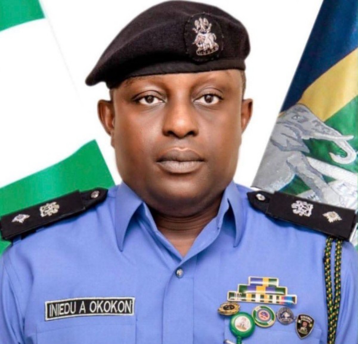 Portrait of a male police officer in a blue uniform with a beret, name tag reads INEDU A OKOKON, medals on his chest, flags in the background.